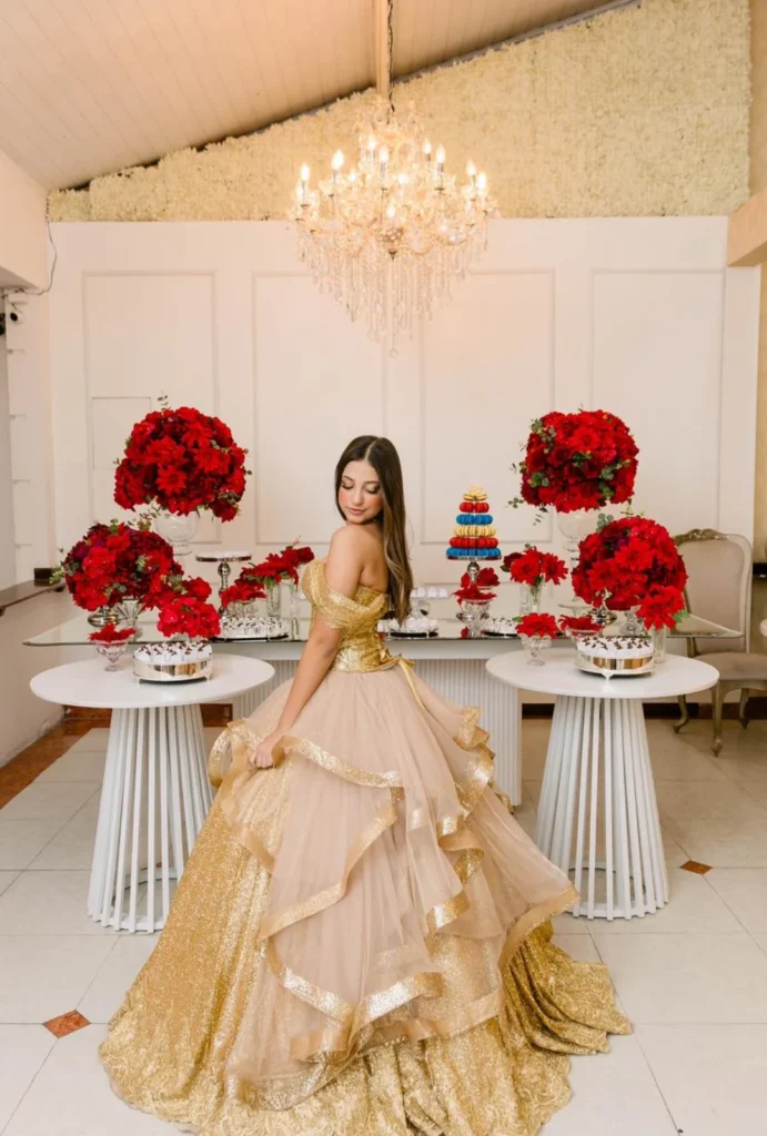 Debutante de vestido dourado ao lado de mesa de doces decorada com rosas vermelhas